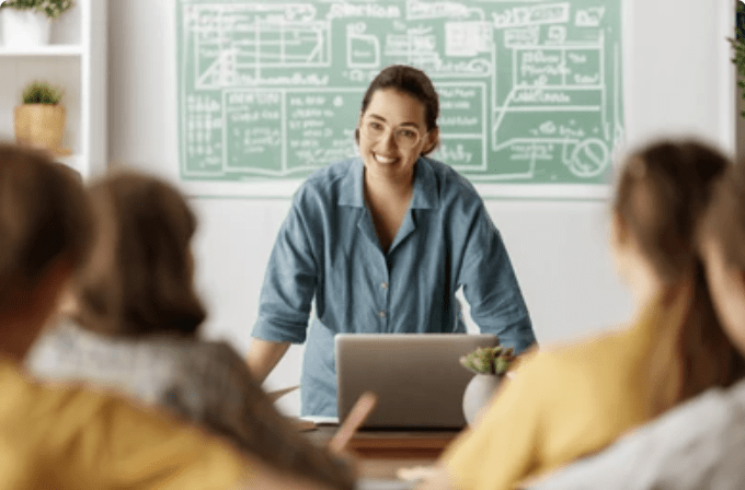 A teacher stands at her desk, behind her computer, and asks her students a question.