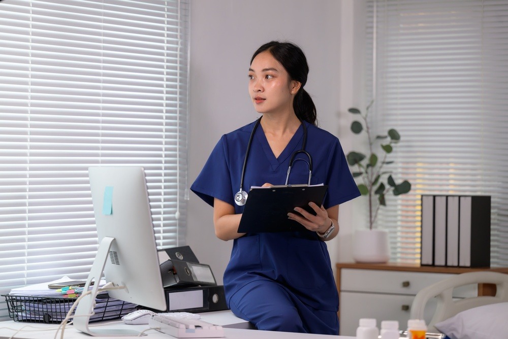 Asian female nurse wearing scrubs and stethoscope, writing on patient chart in a medical clinic