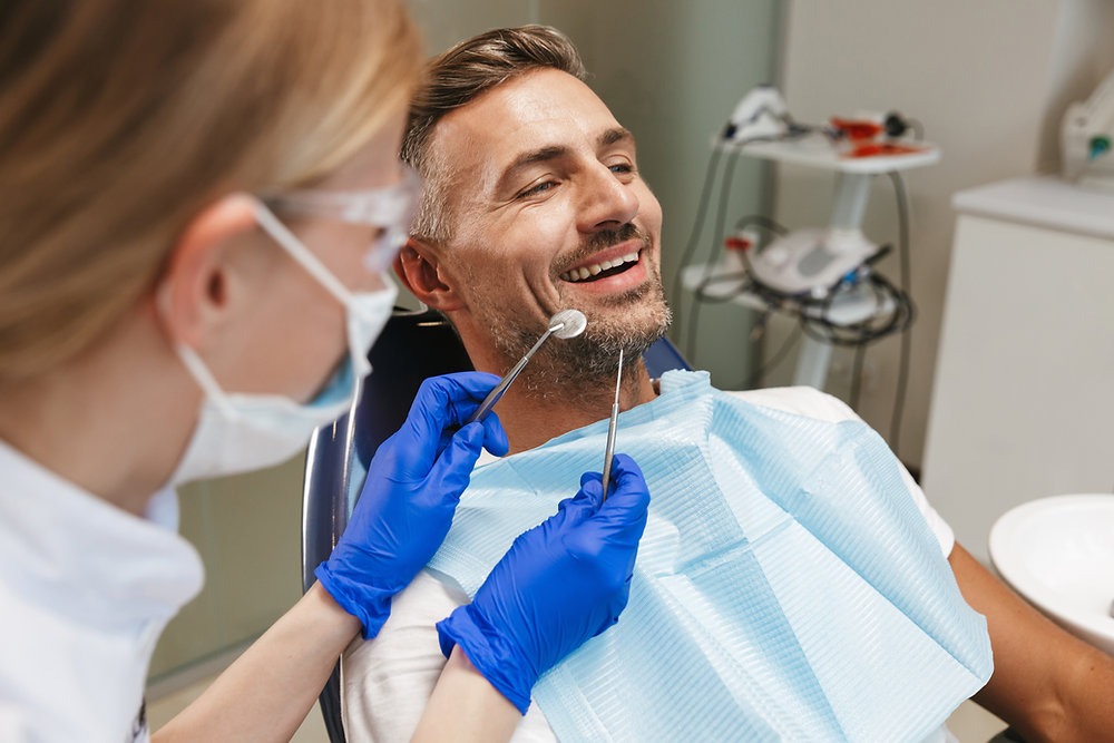 Smiling female dentist wearing blue gloves is examining the teeth of a male patient